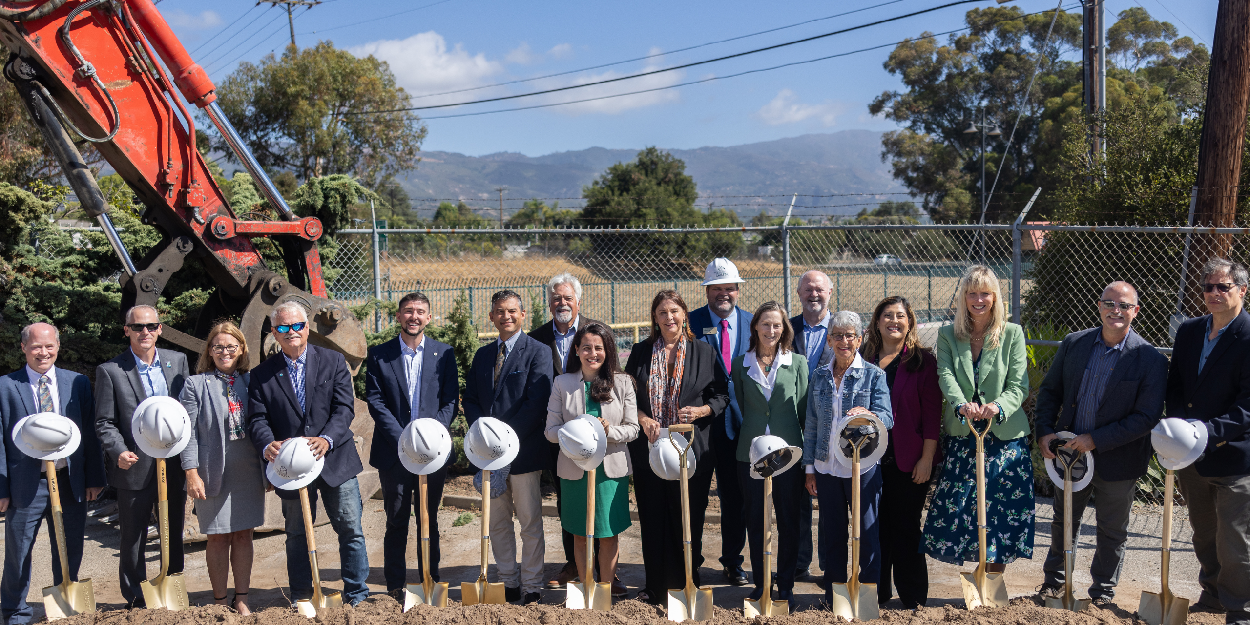 Image of a group of officials from the City of Goleta, State of California and SBCAG behind golden shovels, hardhats and in front of a excavator for the Goleta Train Depot groundbreaking.